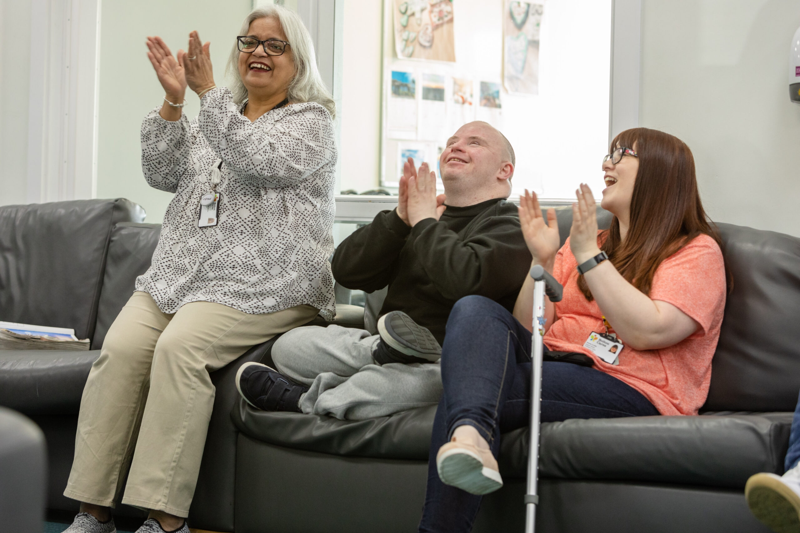 A carer with two young people, they are sitting on a sofa, laughing and clapping