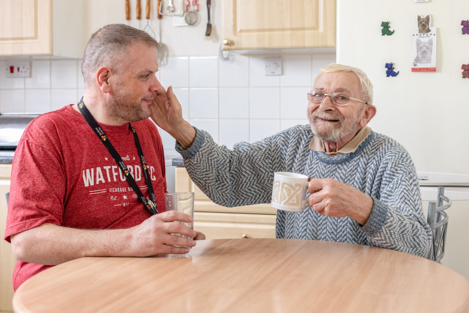 Elderly man having a cup of tea with a volunteer, they are both smiling and the elderly man is patting the carers face gently