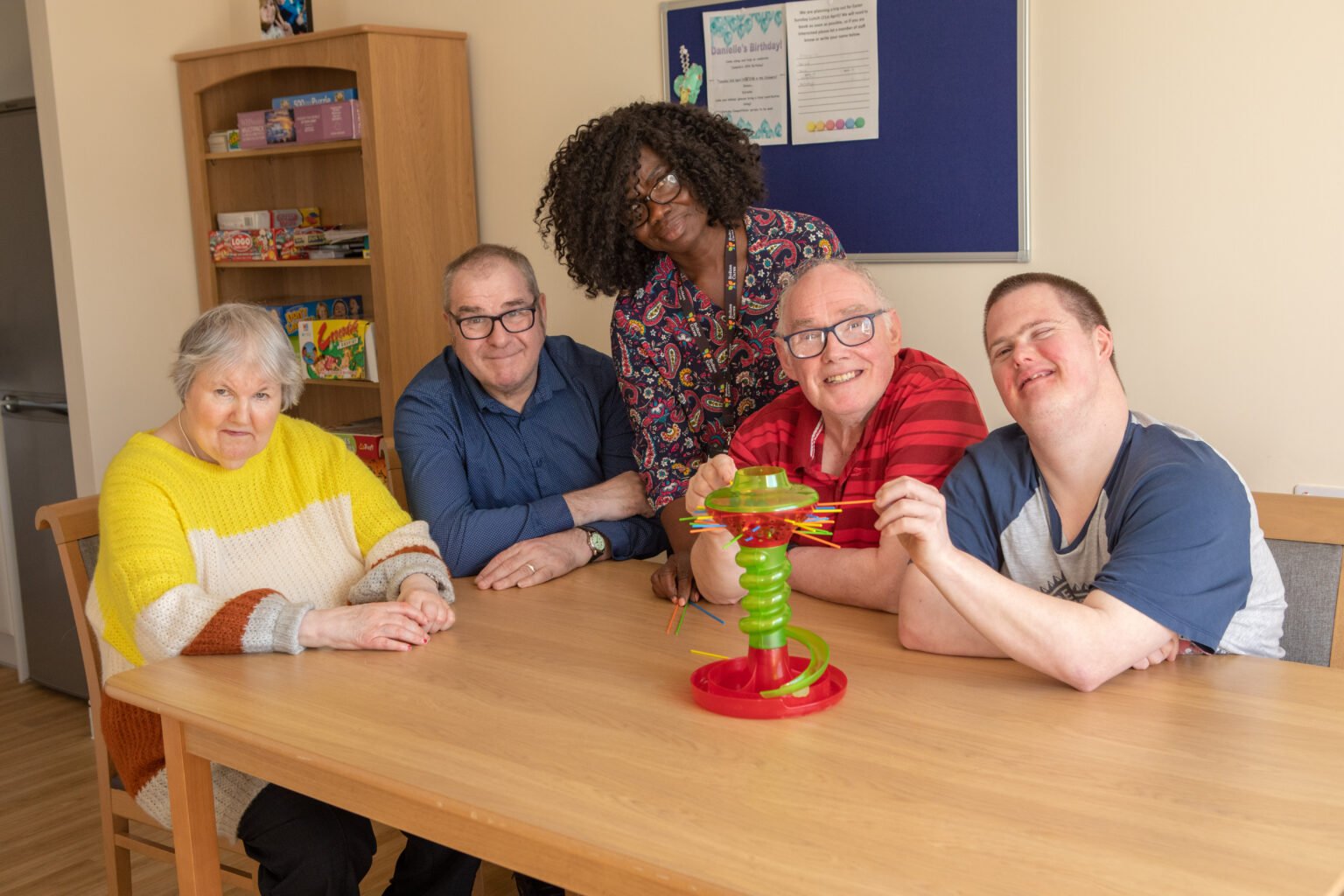 a group of people playing ker-plunk around a table. They are all smiling at the camera