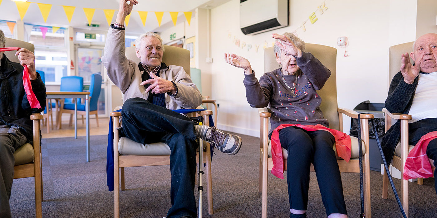 Elderly people sat in a circle waving their arms as they sing along to a song together