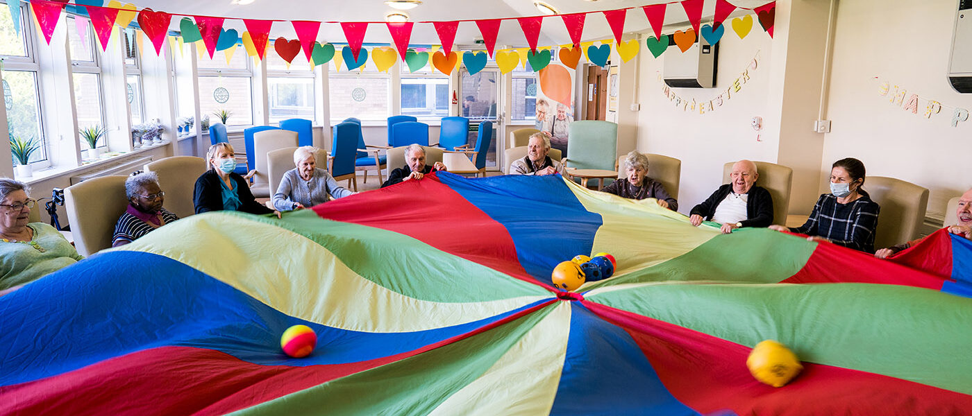 A large parachute is open in the centre of a room - there are people sat in a big circle around it, trying to move soft balls around as a game