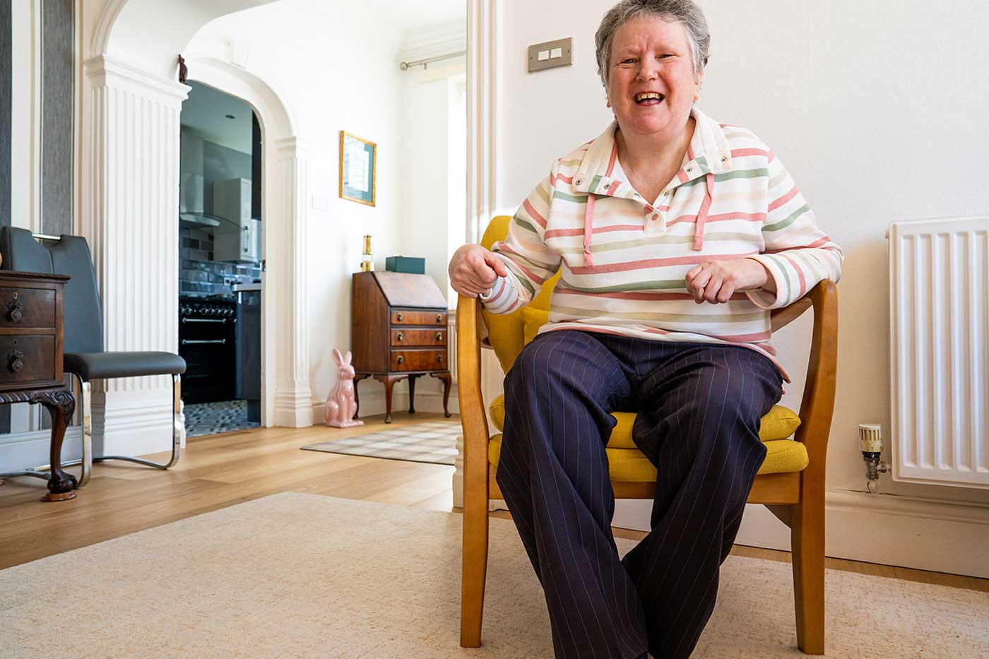 Lady in her living room, she is sat in a chair and smiling at the camera